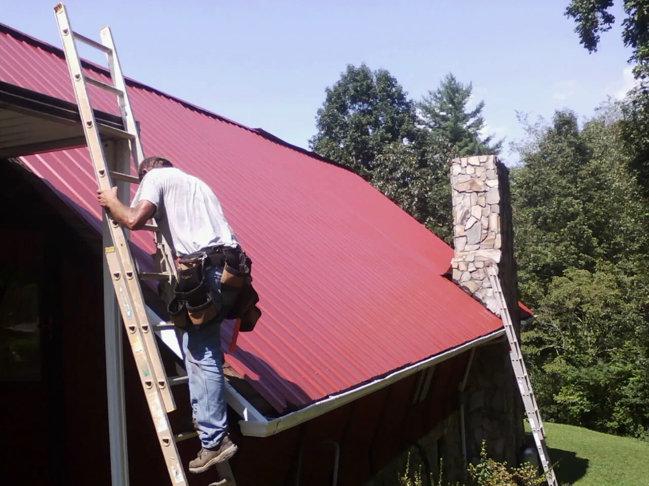 Roofer on a ladder beside a red metal roof and stone chimney — Soffit & Fascia Repair by Roofing Brothers in Spring Mills